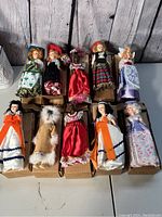 Full top-down photo showing all 10 dolls arranged in two rows inside individual boxes on a light surface, dressed in different traditional costumes, some with hats and colorful fabrics.