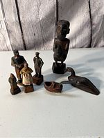 Photo showing seven hand carved vintage items including human figures from Africa and Indonesia and two animal figures plus a carved wood pipe on white surface with wood background