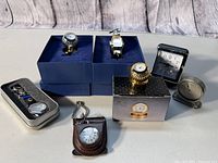 Photo taken showing all items in lot, including two golf ball-themed table clocks, watch-shaped clock, two compact travel alarm clocks, stopwatch in tin, and clock keychain in leather case.