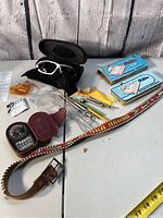 Photo of various vintage items laid out including sports sunglasses with case, leather compass, beaded leather belt, vintage tins, small tools, and a yellow utility knife.