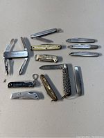 Collection of eleven pocket cutters and small folding knives arranged on a flat surface. Various sizes, some with blades open or closed. Includes two small identical metal cutters with "Sammons Preston" engraving and others with plain metal or white and brass handles.