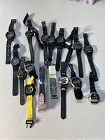 Lot of 13 sport watches of varying styles and colors displayed on a white surface, including digital and analog displays, black, green, yellow, and camouflage straps.
