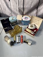 Full view of lot showing assorted vintage vanity items on wood backdrop including powder boxes, electric razor, brush, cologne, lipsticks, and lighter.