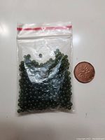 Small clear plastic resealable bag filled with tiny round dark green jade beads. A coin is placed next to the bag for size reference.