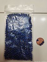 Clear plastic bag filled with numerous small round deep blue lapis lazuli beads next to a penny for scale.
