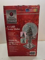 Photo of the boxed Home Accents Holiday 6-inch LED Snow Globe with branding, dimensions, LED feature, and Christmas tree image on packaging.