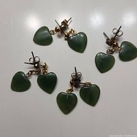 Four pairs of earrings with dark green jade heart-shaped stones attached to gold-tone small balls and silver-tone posts, displayed on white background.