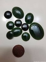 Top view of ten polished dark green jade stones arranged with a coin for scale, showing different shapes and sizes, smooth glossy surfaces that reflect light.