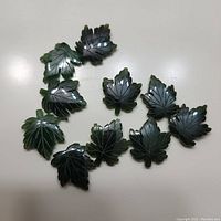 Top-down view of all 10 jade maple leaf carvings showing their dark green color and polished surface.