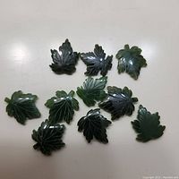 Top view photo showing all 10 dark green jade maple leaf pendants arranged on a white surface. The carvings and vein details are visible on polished jade.