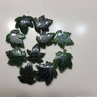 Top-down photo of 10 dark green jade maple leaf carvings arranged in a circle, showing engraved vein details and polished surfaces.