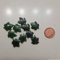 View of 10 small dark green jade maple leaf carvings next to a Canadian penny for size reference.