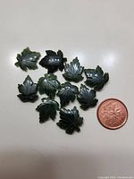 Top view photo showing 10 green jade maple leaf carvings arranged with a one-cent coin for size comparison on a white surface.