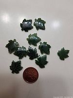 Top view of ten dark green jade maple leaf carvings arranged on a white surface with a penny for scale