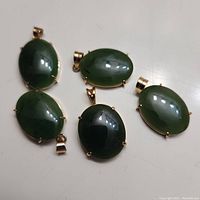 Five green jade oval pendants with gold-tone bails arranged on a white surface showing top view and side details.
