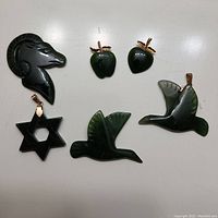 Top-down view of 6 carved dark green jade pendants arranged on white background showing details of shapes and gold-colored bails