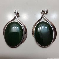 Two oval dark green jade pendants set in silver-tone metal frames with leaf-like design sides, shown from front