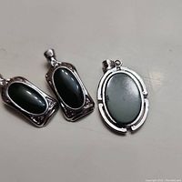 Three jade pendants: two dark green oval stones in rectangular silver-tone frames and one lighter green oval stone in an oval silver-tone frame.