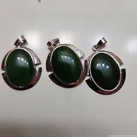 Three oval green jade pendants with silver-tone metal frames, showing front view and bail for chain attachment.