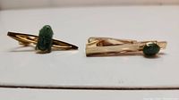 Two gold-tone tie clips displayed on a white surface, one with a carved jade figure and one with an oval jade stone.