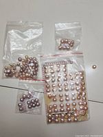 Multiple bags of pearls in various pink, white, and peach tones for jewelry making, arranged on a flat surface.