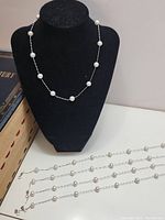View of five white pearl necklaces displayed with one on a black necklace stand and four lying flat. Pearls are spaced evenly on thin silver-color chains.