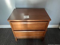 Front view of mid century modern walnut nightstand with two drawers against a light blue wall, showing the overall shape and design, with a tag numbered 4 placed on top.