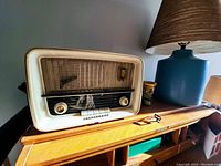 Full view of the Telefunken Jubilee radio showcasing its cream-colored casing with gold-accented tuning knobs, placed on a wooden surface next to a blue lamp.