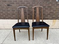 Pair of vintage mid century side chairs with high backs, black leather seats, and walnut stained beechwood frames photographed against brick wall on pavement.