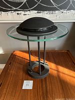 Full view of the lamp showing the glass top, black metal rods, round base with hole, and black dome shade.
