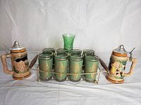 Front view of entire lot showing eight green glasses with gold design in round metal and wood carrier, two ceramic beer mugs with hinged pewter lids, and green vase in background