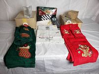 Full lot showing boxed medium and smaller clear triangular vases, two stacks of embroidered Christmas towels in green and red, white folded tablecloths laying flat on grey cover, and additional packaged textiles in the back.