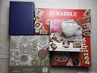 Photo showing Scrabble board game, coffee cup puzzle, Yahtzee game, In Bloom puzzle, and a blue folder or binder on a table.
