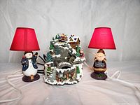 Photo showing whole lot: Christmas fountain decoration centered between a snowman lamp and a Santa lamp, both with red lampshades.