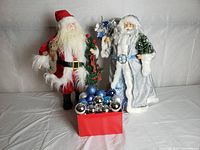 Two Santa Claus figures standing side by side with a red box filled with blue and silver Christmas tree balls in front of them. The figure on the left in a red suit, the figure on the right in a silver/blue coat.