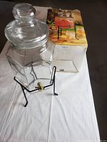 Glass decanter with lid and brass spigot placed on a black metal stand next to its original box with visible damage.