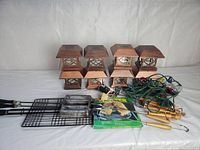 Eight solar pole lights stacked 2 high in 4 columns with solar panels visible on top. In front left rectangular wire mesh BBQ baskets with handles. Center front multi-colored Christmas bulbs on green wires. Front right metal skewers with wooden handles.