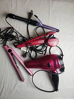 Three hair care tools laid out on a white surface: pink Revlon hair dryer, pink flat iron, and purple curling iron with cords visible.
