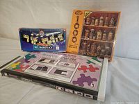 Photo showing the unopened Rummy-O tile game, 1000-piece jigsaw puzzle box depicting collectible mugs, and puzzle tote in new condition.