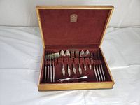 Closed wooden case with brown velvet interior holding silver plate flatware pieces including knives, forks, spoons, and serving utensils organized in slots.