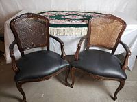 Front view of two vintage wooden armchairs with carved details, cane backs, and black leather seats.