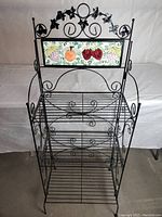 Front view of black wrought iron kitchen stand with three wire shelves and decorative fruit tile panel.