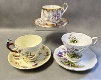 Three bone china teacup and saucer sets arranged on neutral backdrop: pink rose and gold trim (Stratford) at top, bird-branch handle with colorful birds (Grafton) at left, purple violets with gold edging (Juliet) at right.
