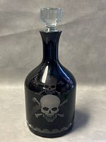 Full front view showing the black glass decanter with etched skull and crossbones design and attached clear glass stopper on top.