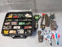 Opened black plastic case with compartments holding various military model toys including planes, tanks, vehicles, artillery, and accessories.