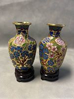 Pair of Chinese cloisonné bud vases with floral enamel patterns on dark wood bases, shown standing side by side on a neutral background.
