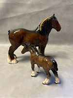 Two porcelain figurines of a large Clydesdale horse and a smaller colt viewed front and side.