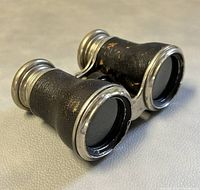 Angled side view of the vintage LE JOCKEY CLUB Paris binoculars, showing textured black body and metal rims with wear and peeling.