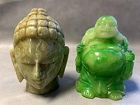 Front view of vintage onyx stone Buddha head with serene facial features beside green resin happy Buddha figurine showing detailed carving and glossy finish.