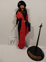 Full view of Barbie doll wearing black dress, red shawl, black hat, and standing next to black display stand.